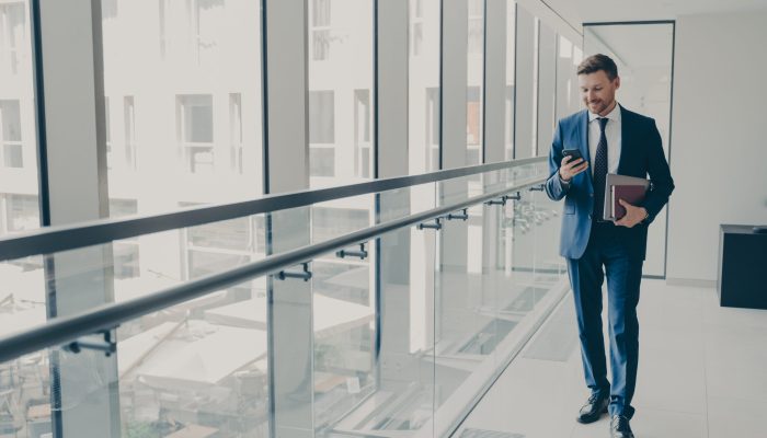 Smiling redhead man office worker in suit using cellphone, checking email or news feed of social network account with laptop and notebook in hand, looking at smartphone while standing in office